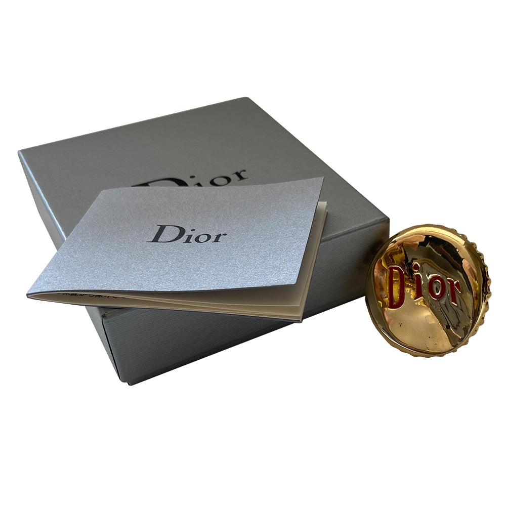Christian Dior Logo Bottle Cap ring circa 2004. Gold-tone "bent” bottle cap ring with red embossed logo and thick band. Adjustable band for various sizing. Dior box and booklet included.