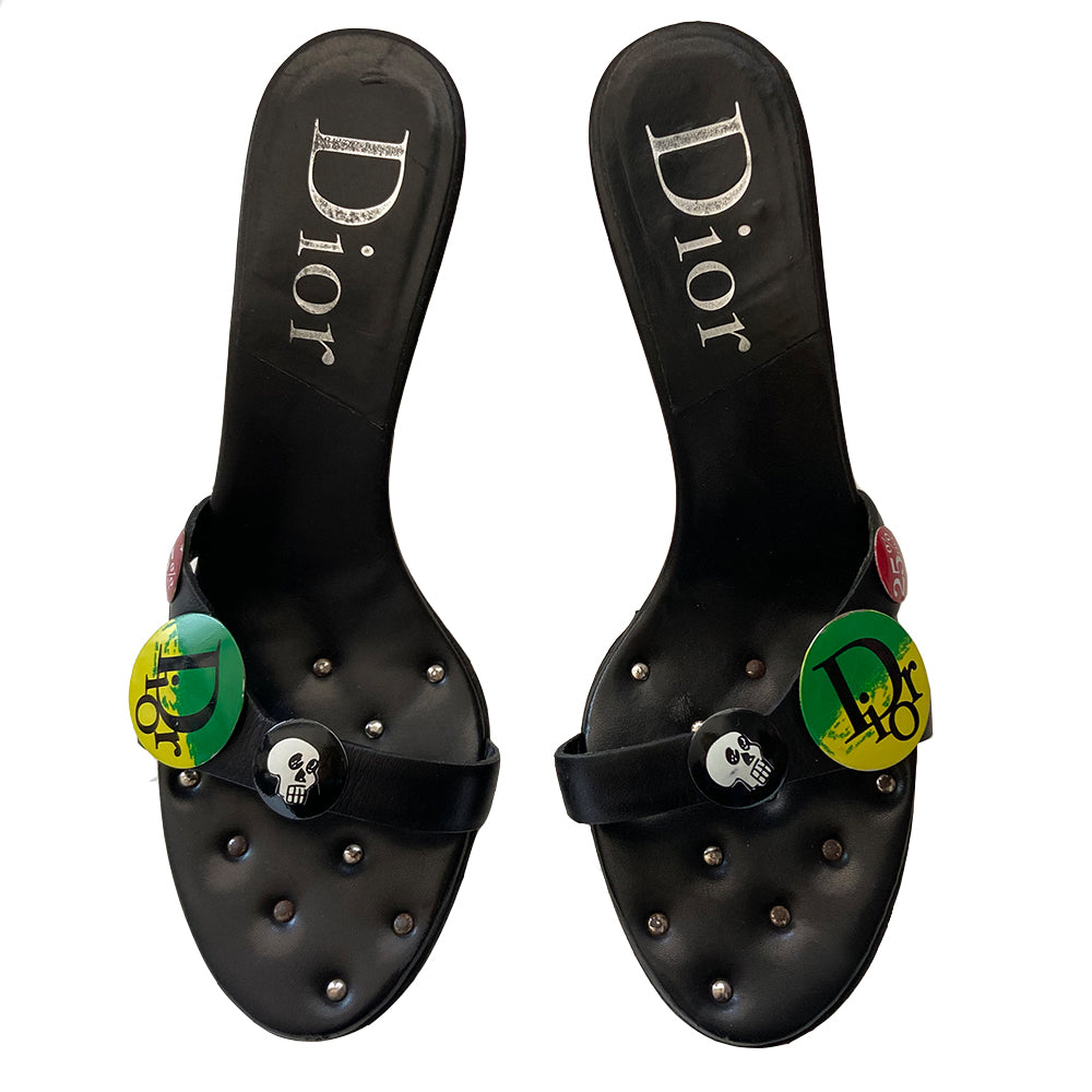 Christian Dior black leather slip-on open toe sandals with silver-tone metal high heels, John Galliano for Dior Spring 2003 with skull, Dior logo and 25% off badges attached to upper leather straps. Leather with silver-tone stud tufting inside front insole. Black with green, yellow, red, white buttons. Made in Italy