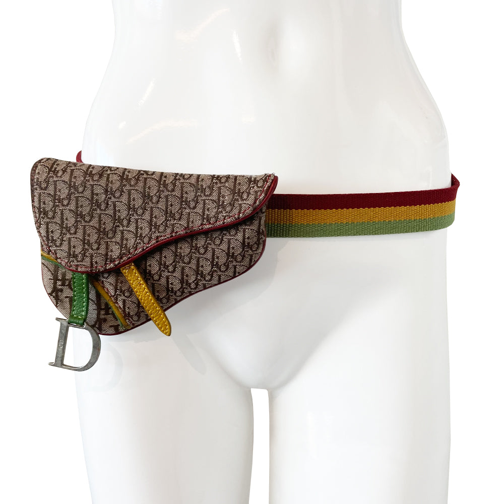 Christian Dior Rasta mini saddle belt waist bag by John Galliano for Dior 2004. Diorissimo canvas with red leather outer piping, yellow & green accent piping, silver-tone hardware. Adjustable canvas webbing & leather belt with 5 hole pin closure. Top flap magnetic snap closure with green & yellow leather straps, CD engraved hanging D, brown satin textile interior.